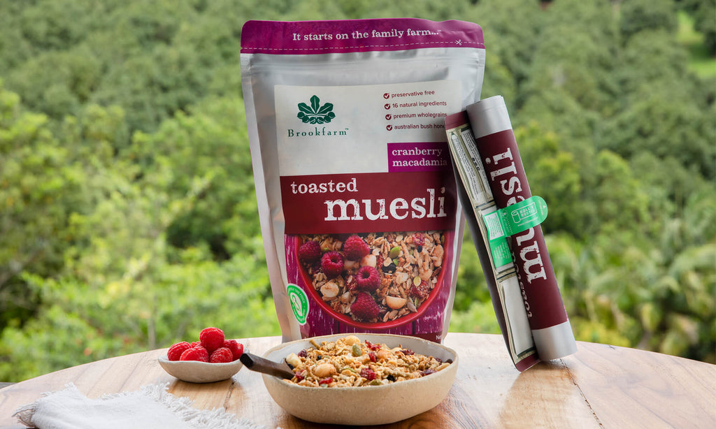 Roll'n'recycle packaging stands behind bowl of toasted muesli with raspberry in a ceramic bowl. Macadamia orchard in the background